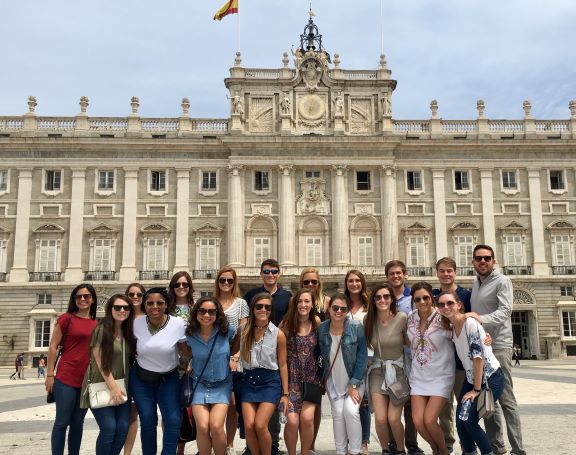 Goup of students in front of a government building in Madrid. They are crowded together to fit in the frame. It is a sunny day and all are smiling.