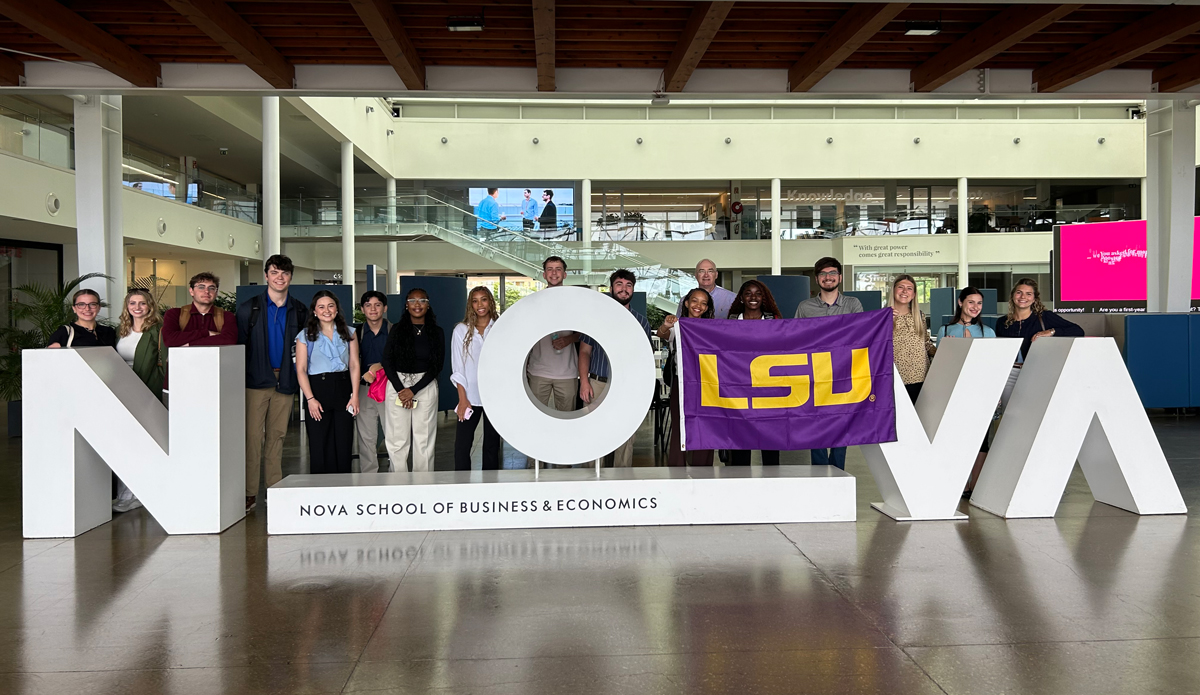 More than a dozen LSU students pose outside the entrance of the NOVA School of Business & Economics in Portugal. They stand amongst large white letters spelling out NOVA. Three students hold a purple and gold LSU flag.