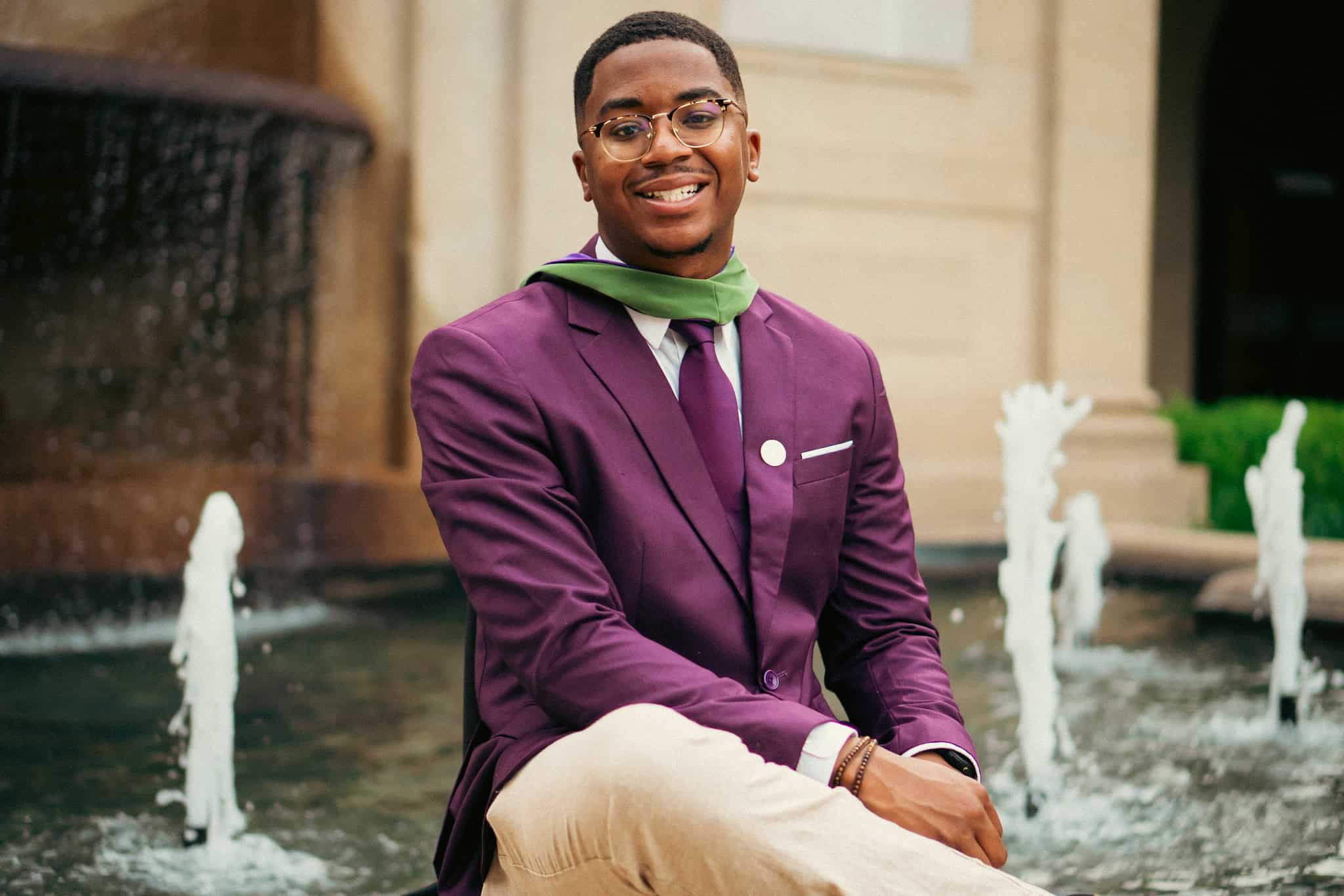 Jared Smith wearing a purple suit jacket with a Masters graduate hood sitting in front of the Dodson Hall fountain on LSU campus